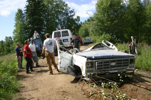 2003 Jeep TJ - A group of people stand around a heavily damaged white pickup truck on a dirt road, with other vehicles visible in the background.