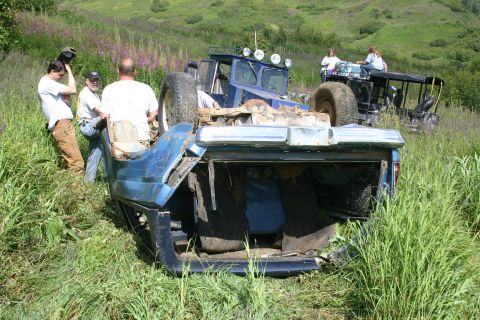 2003 Jeep TJ - An overturned blue pickup truck lies in a grassy field, with several people standing around and another vehicle visible in the distance.