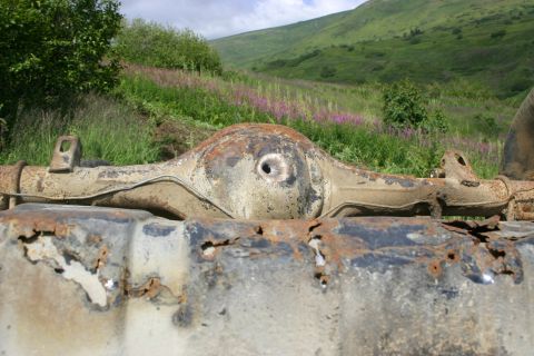 2003 Jeep TJ - A heavily rusted rear axle differential is visible in the foreground, with a hole in its center. The background shows a green, grassy hills…