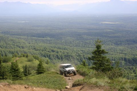 2003 Jeep TJ - A white Jeep Wrangler navigates a steep, dusty trail amidst a lush green landscape with distant mountains.