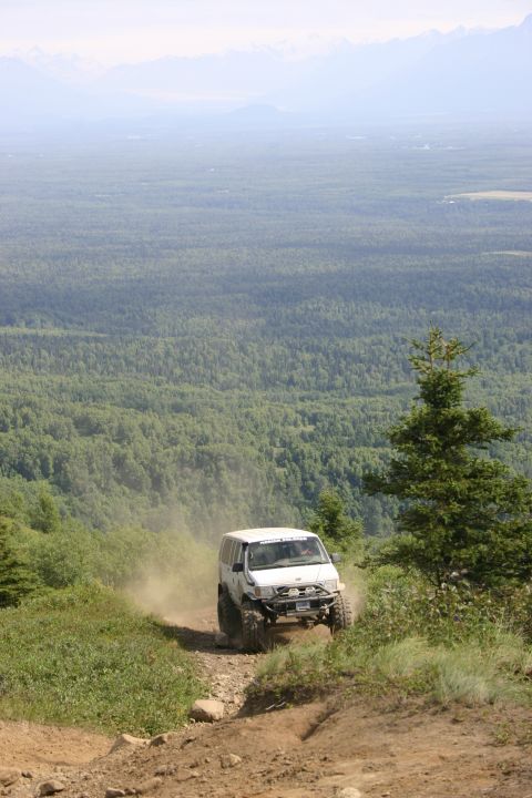 2003 Jeep TJ - A white 2003 Jeep TJ navigates a steep, dusty trail with a vast forest and mountains in the background.