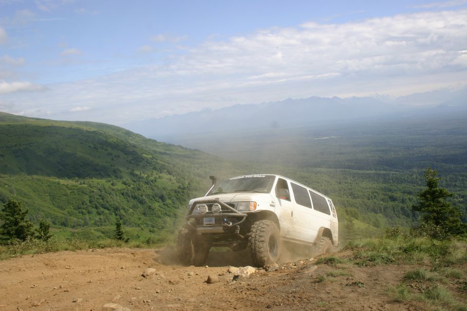 White van with big tires kicking up dust on a dirt road with a mountain view