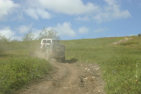 2003 Jeep TJ - Two off-road vehicles navigate a dusty dirt path through a grassy landscape under a partly cloudy sky.