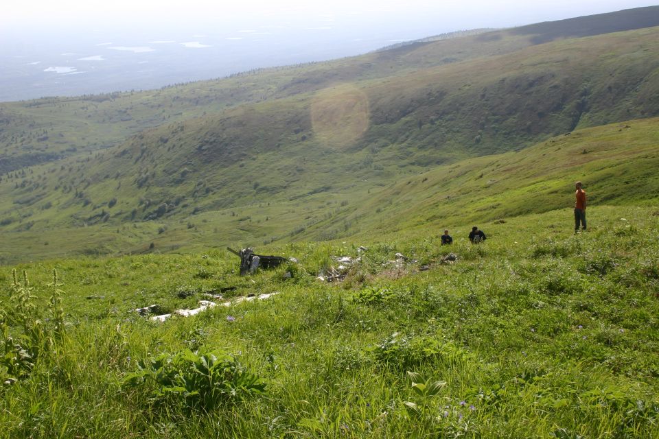 Several people stand on a grassy mountainside with a distant view of lakes