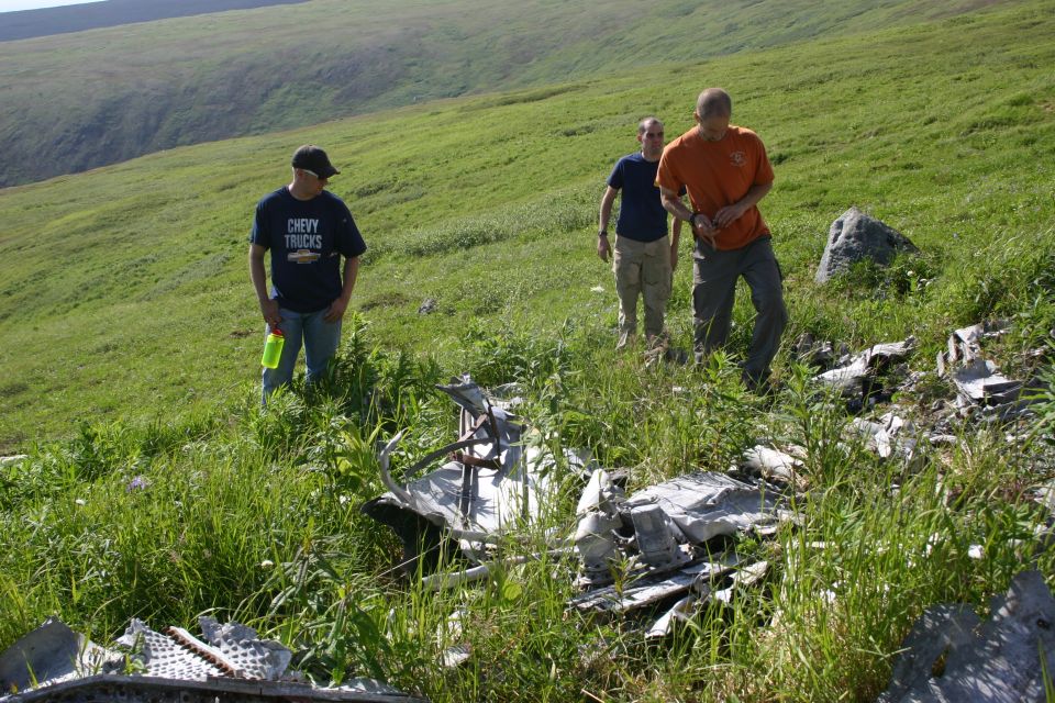 three people looking at plane wreckage scattered in tall grass on a hillside