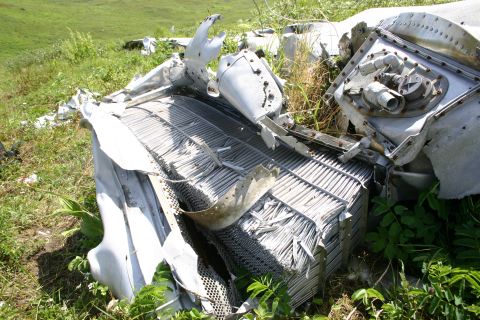 2003 Jeep TJ - Debris from an aircraft crash lies scattered in a field, with green grass and foliage surrounding the mangled metal.