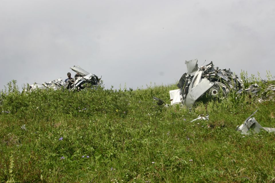 Plane wreckage scattered on a grassy hillside under a cloudy sky. Two people are visible near the debris on the left