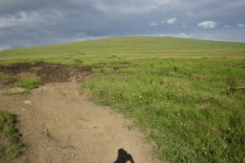 2003 Jeep TJ - A dirt path winds through a grassy field towards a large, green hill under a dramatic, cloudy sky.