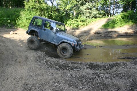 2003 Jeep TJ - A blue 2003 Jeep TJ navigates a muddy off-road course, with one wheel submerged in a water puddle.