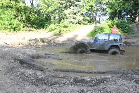 2003 Jeep TJ - A blue Jeep Wrangler navigates a muddy obstacle course, kicking up water and mud.