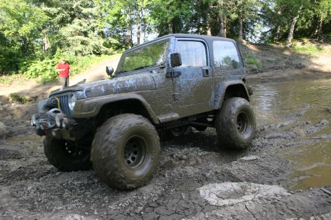 2003 Jeep TJ - A blue Jeep Wrangler navigates a muddy off-road course, splashing through water and mud.
