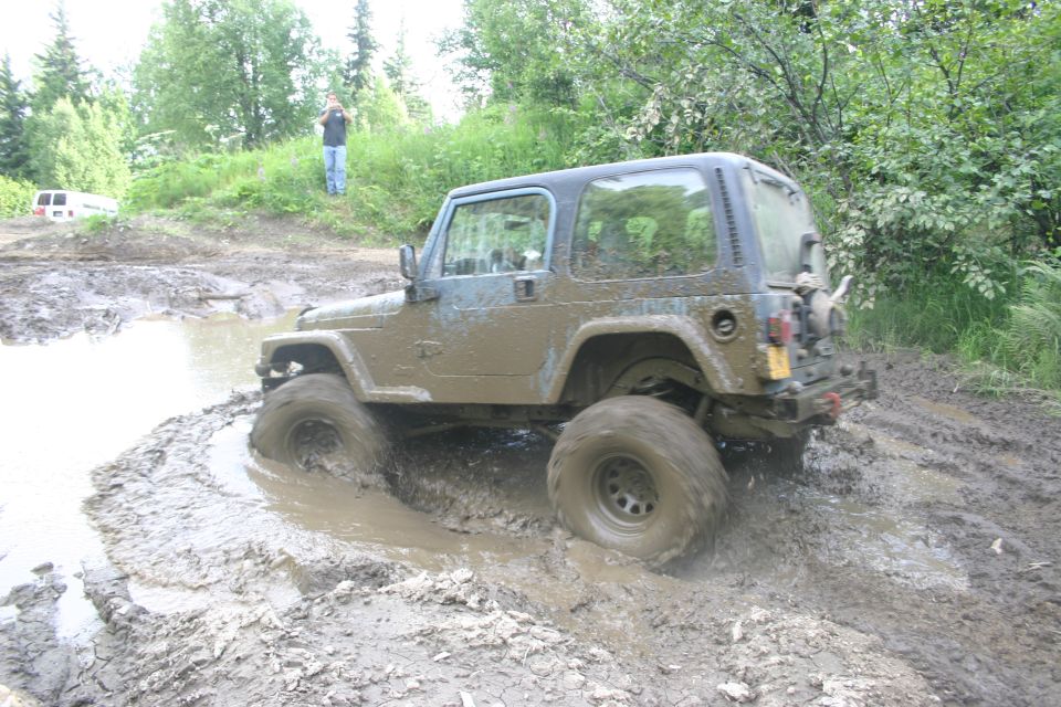 2003 Jeep TJ struggling through deep mud on an off-road trail. Another white van is visible in the background