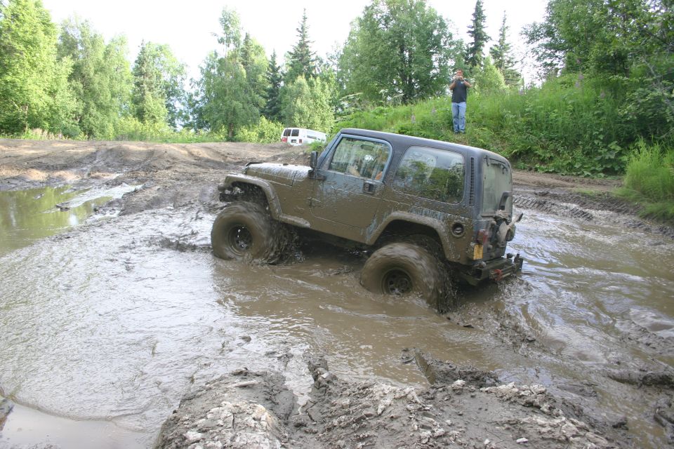 Muddy Jeep TJ driving through a deep mud pit with a white van in the background