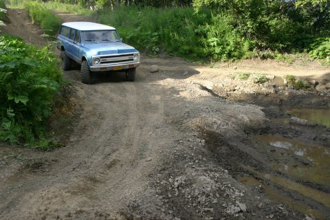 2003 Jeep TJ - A blue Chevrolet Suburban navigates a muddy, off-road trail with dense foliage on either side.