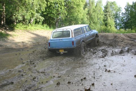2003 Jeep TJ - A blue and white long wheelbase SUV navigates a muddy off-road course, kicking up spray as it goes.