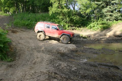 2003 Jeep TJ - A red Jeep Cherokee XJ navigates a muddy off-road course with trees in the background.