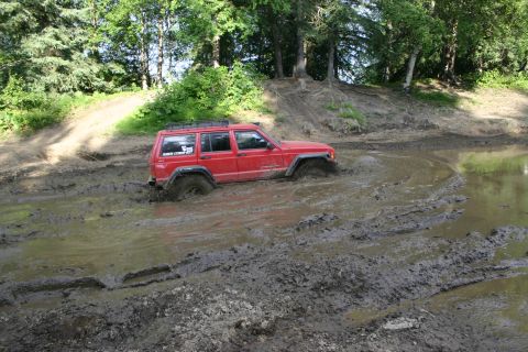 2003 Jeep TJ - A red Jeep Cherokee navigates through a muddy terrain, with its wheels submerged in thick mud.