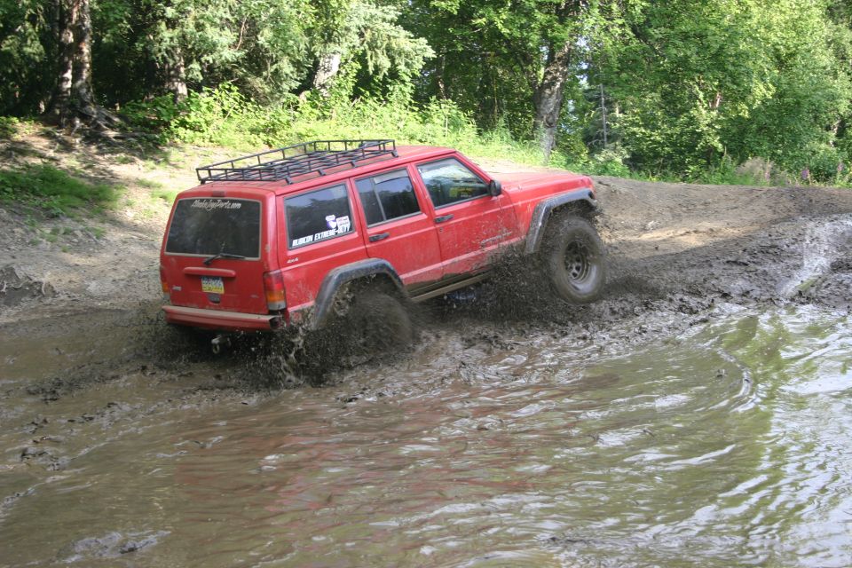 Red Jeep Cherokee XJ with a roof rack driving through a muddy puddle, kicking up water