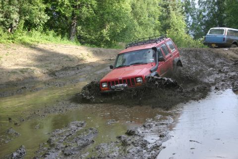 2003 Jeep TJ - A red Jeep Cherokee XJ navigates a muddy trail, kicking up water and dirt.