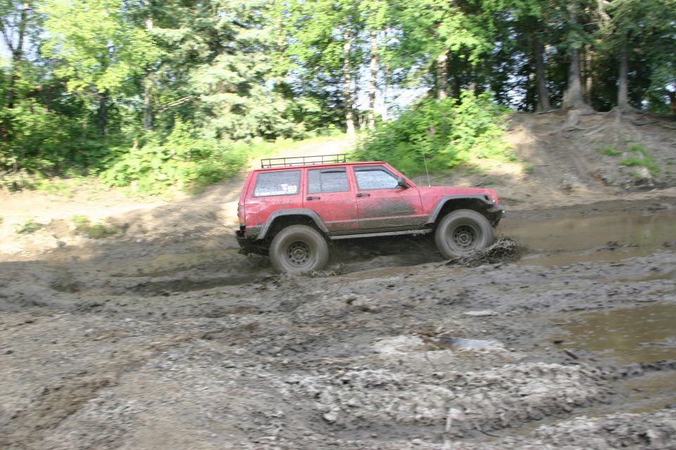 Red Jeep Cherokee XJ with roof rack driving through a muddy trail, kicking up mud