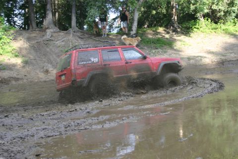 2003 Jeep TJ - A red Jeep Cherokee XJ navigates a muddy off-road course, kicking up water and dirt.