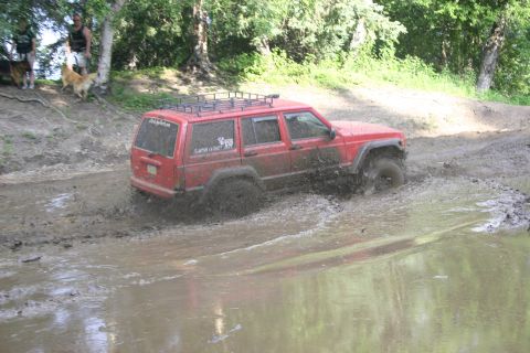 2003 Jeep TJ - A red Jeep Cherokee XJ navigates a muddy trail, kicking up water and mud as it drives through a puddle.