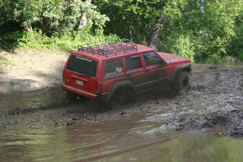 2003 Jeep TJ - Red Jeep Cherokee XJ navigates a muddy trail, kicking up water and dirt.