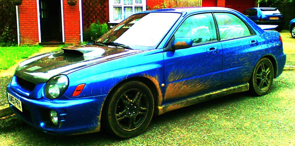 Blue Subaru Impreza sedan with a hood scoop and black wheels, covered in mud, parked on a street