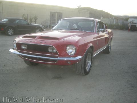 1990 Ford Bronco - red Shelby Mustang fastback is shown front three-quarter on gravel with its Shelby hood scoop and grille visible.