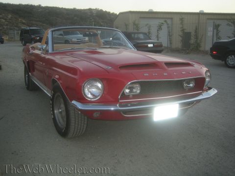 1990 Ford Bronco - red Shelby Mustang convertible faces the camera with one headlight glowing, parked on gravel near a building.