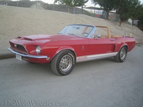 1990 Ford Bronco - red Shelby GT500KR convertible with side stripes sits front three-quarter on gravel beside a dirt mound.