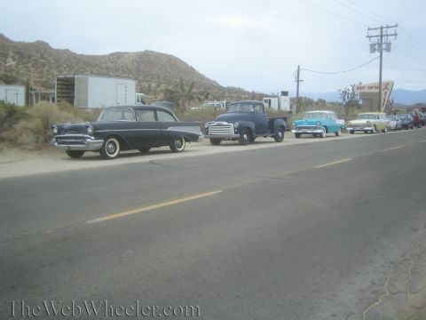 1990 Ford Bronco - line of vintage 1950s cars and a pickup truck parked along a rural desert road under an overcast sky.