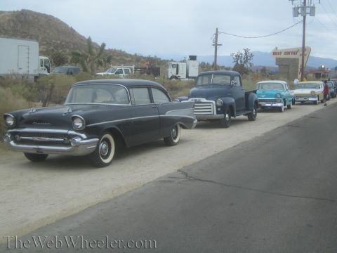 1990 Ford Bronco - dark 1950s Chevrolet sedan parked at the curb leading a row of vintage cars and a pickup along a desert road.
