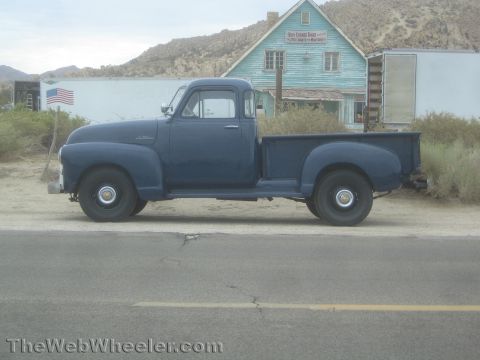1990 Ford Bronco - Side profile of a dark blue 1950s pickup truck with a step-side bed parked along a desert road by a weathered building.