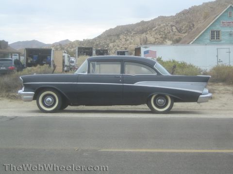 1990 Ford Bronco - Side profile of a black 1957 Chevrolet two-door sedan with chrome trim and whitewall tires parked on a desert road.
