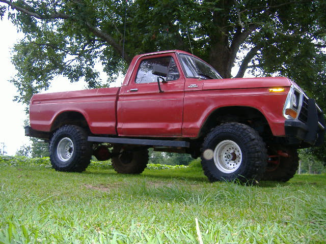 Red 1979 Ford F-150 pickup truck with large tires and a brush guard, parked on grass under a tree