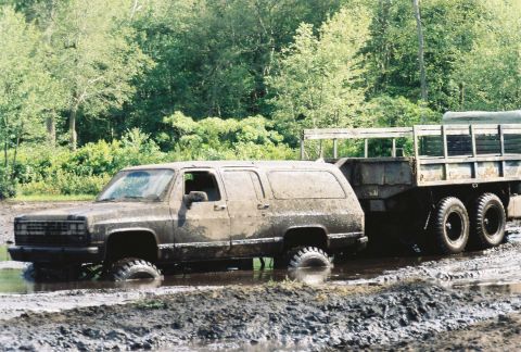 1991 Chevrolet Suburban - A muddy SUV tows a trailer through a challenging off-road course, with trees in the background.