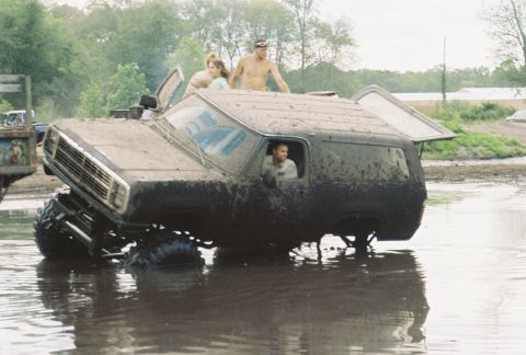 1991 Chevrolet Suburban - A heavily modified SUV is stuck in a muddy body of water with people riding on its roof and inside.