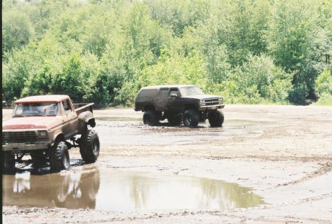 1991 Chevrolet Suburban - Two modified 4x4 trucks navigate a muddy off-road course with trees in the background.