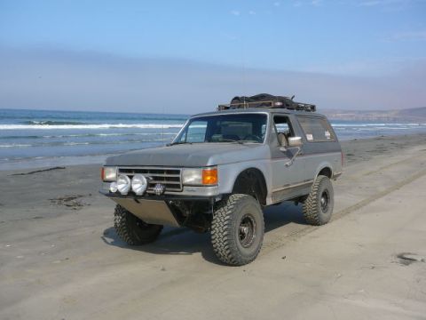 1988 Ford Bronco - Lifted silver 1987-91 Ford Bronco 4x4 with brush guard, off-road lights and roof rack parked on a sandy beach.