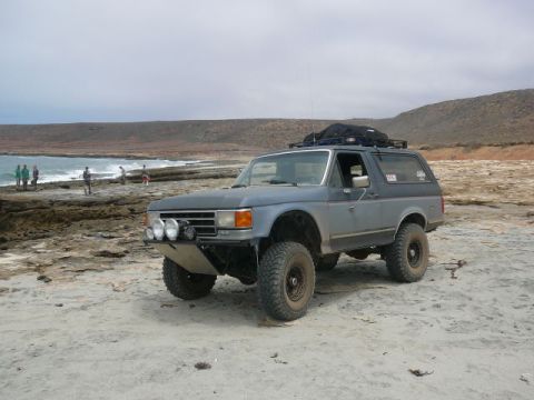 1988 Ford Bronco - Lifted primer-gray full-size Ford Bronco parked on a sandy beach beside a rocky coastal shelf, shot from a low 3/4 front angle. Build detai…