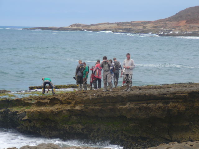 Group in riding gear standing on rocky coastal shelf above breaking surf