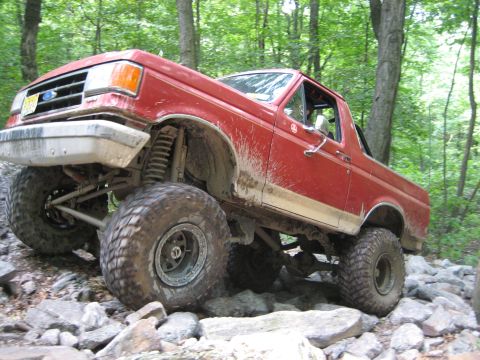 1990 Ford Bronco - Mud-splattered lifted red full-size Ford Bronco crawling over a rock garden on a wooded trail, articulating its front suspension with one w…
