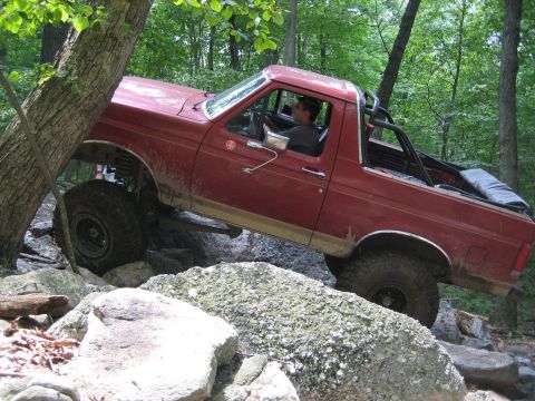 1990 Ford Bronco - Driver-side view of a mud-splattered lifted red Ford Bronco climbing a rocky ledge on a wooded trail with the driver visible behind the whe…