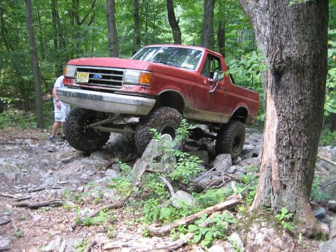 1990 Ford Bronco - A red Ford Bronco is shown off-roading on a rocky, wooded trail.