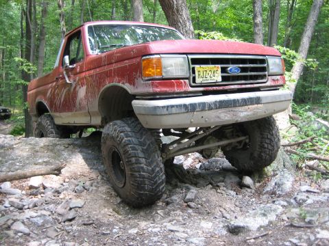 1990 Ford Bronco - Mud-streaked lifted red Ford Bronco with the top removed, posed nose-down on a rocky ledge in the woods at a front three-quarter angle. The…