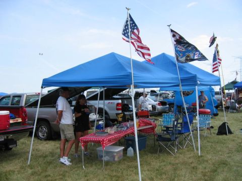 1985 Ford Crown Victoria LTD - People gather under blue tents with American flags at an outdoor event, with several trucks parked nearby.