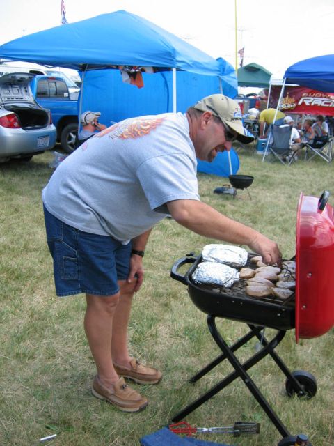 1985 Ford Crown Victoria LTD - A man grills food at an outdoor event with tents and vehicles in the background.