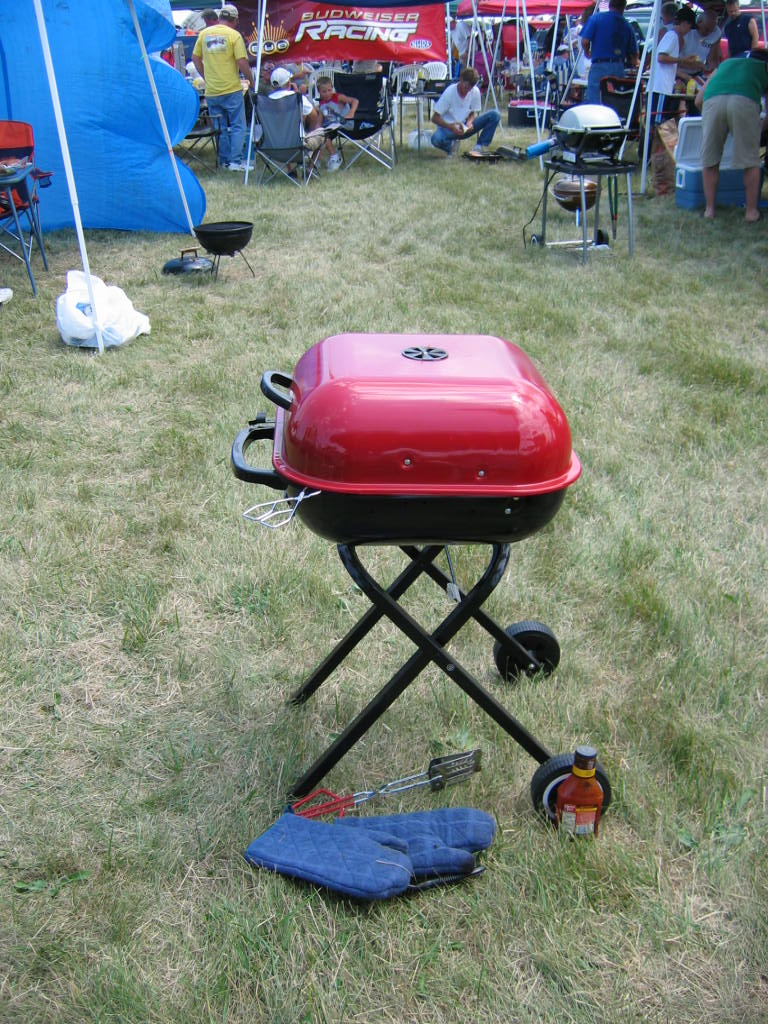 Red portable grill on folding legs with tongs, mitts, and a bottle of sauce on grass