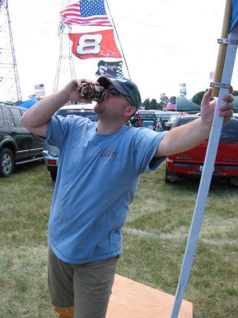 1985 Ford Crown Victoria LTD - A man enjoys a drink at an outdoor event with flags and vehicles visible.
